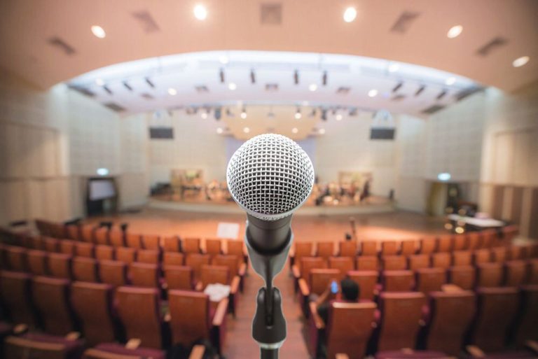 Micrófono en primer plano, con un auditorio vacío al fondo.
